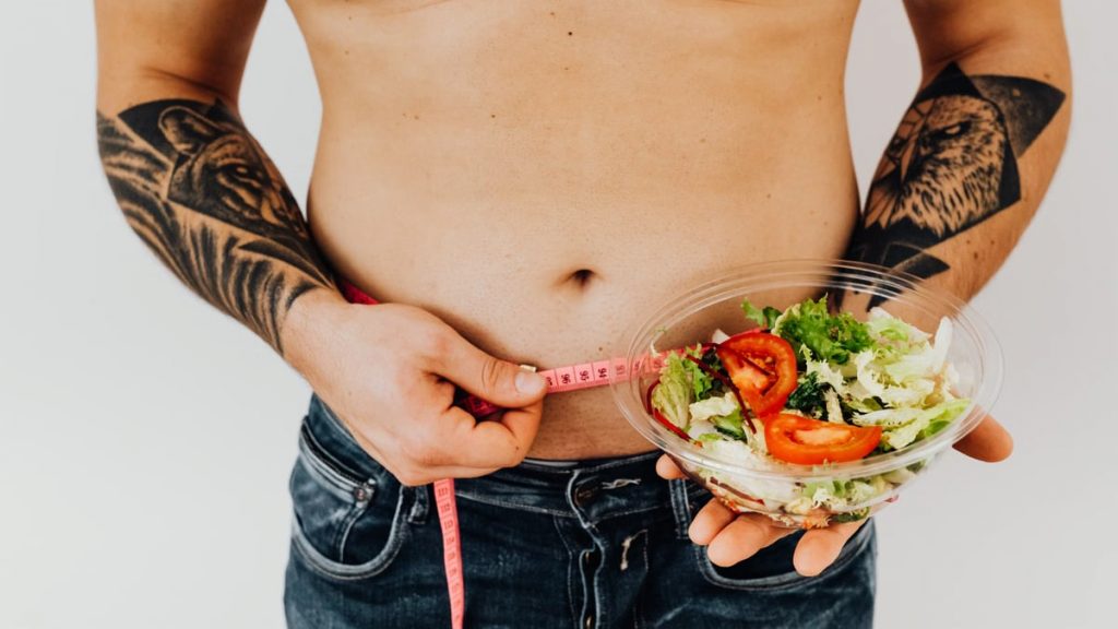 Tattooed man measuring waist while holding fresh salad, symbolizing healthy lifestyle and weight management.