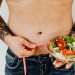 Tattooed man measuring waist while holding fresh salad, symbolizing healthy lifestyle and weight management.
